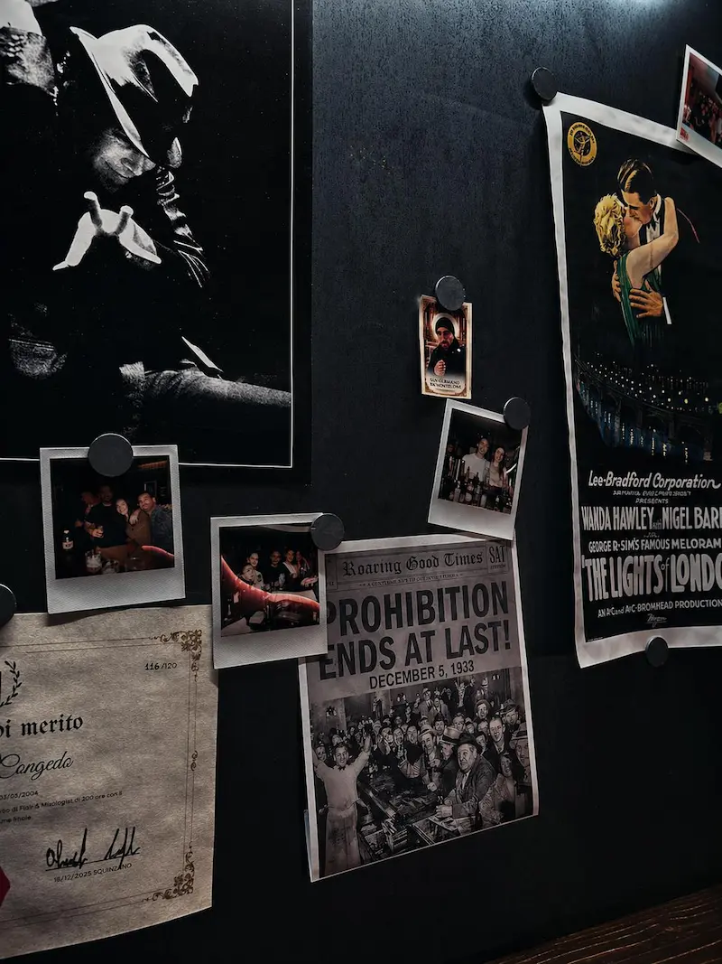 Parete con manifesti d'epoca del Proibizionismo, polaroid e attestato di mixology nel cocktail bar di Lecce
